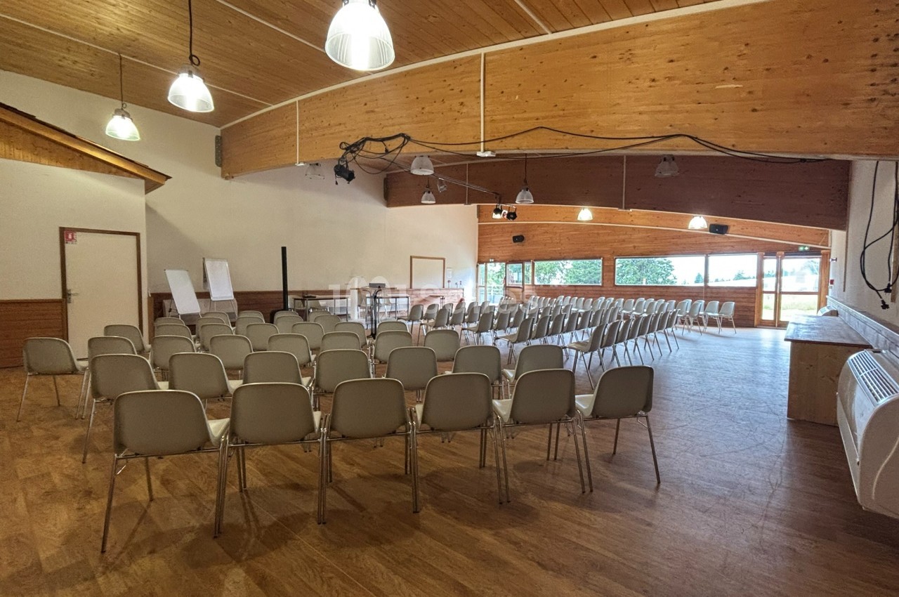 Salle de réunion avec des rangées de chaises alignées, un plafond en bois et de grandes fenêtres laissant entrer la lumière…