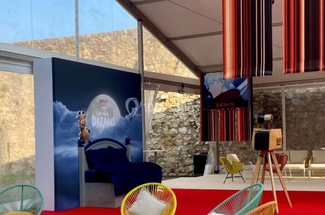 Espace lounge sous une structure transparente, avec fauteuils colorés, tapis rouge et décorations suspendues.