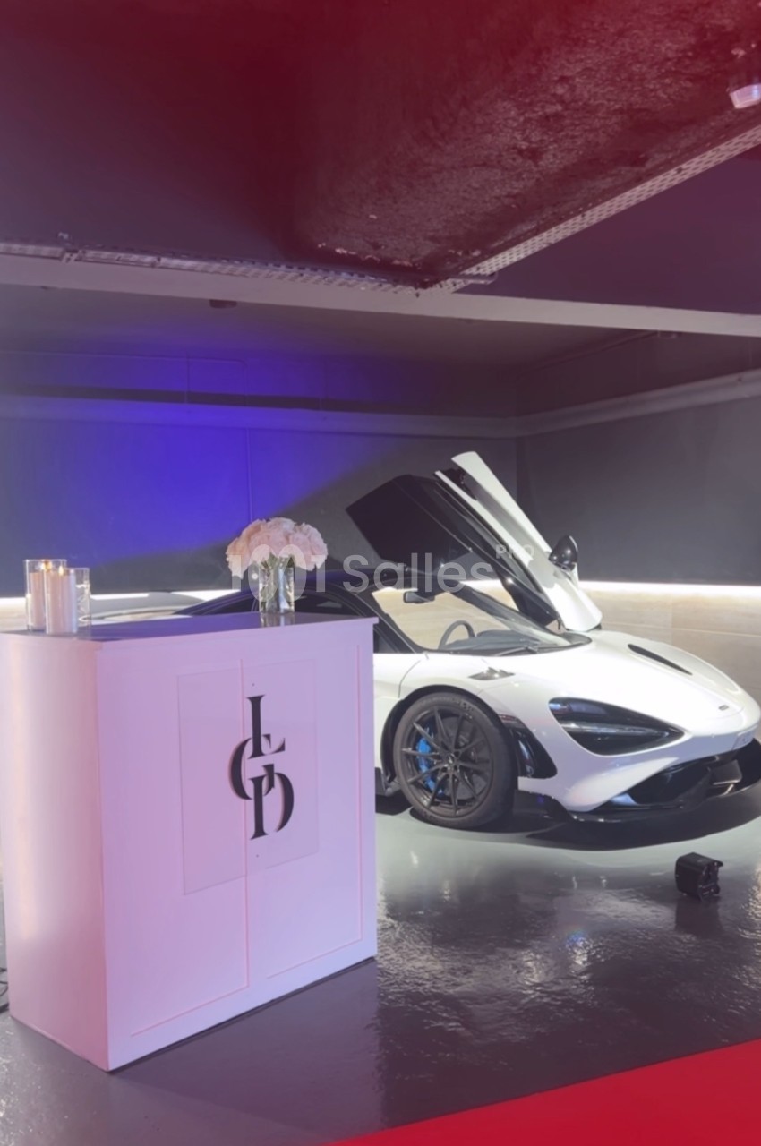 Voiture de sport blanche aux portes ouvertes exposée dans un espace moderne avec éclairage tamisé et décor minimaliste.