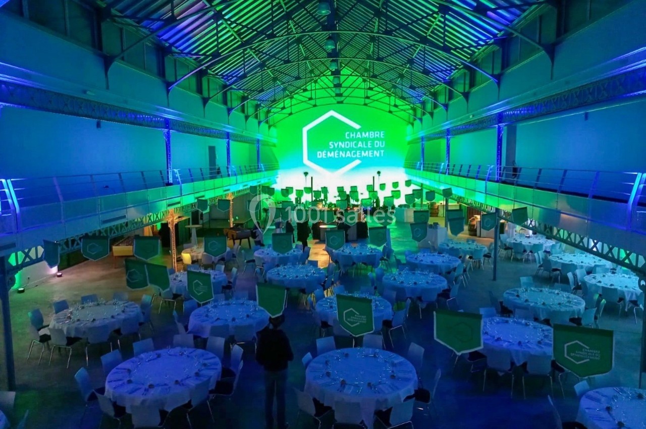 Salle de réception illuminée en vert avec des tables rondes dressées et une scène au fond pour un événement.