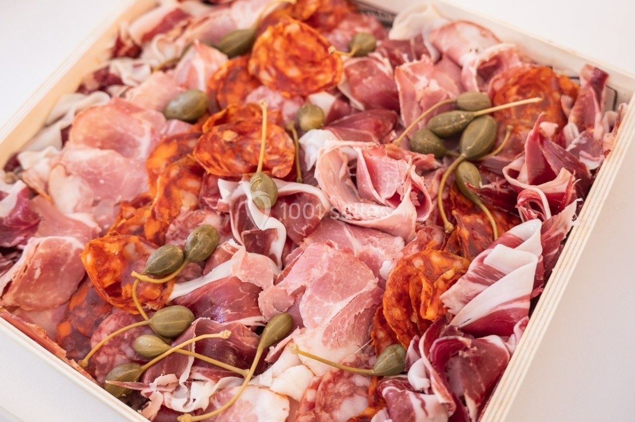 Plateau de charcuterie variée avec jambon, chorizo, caprons et autres viandes froides disposées dans un contenant.
