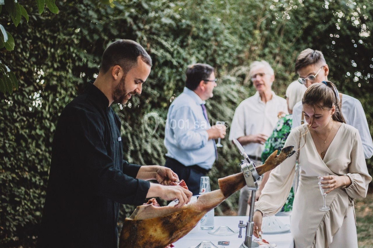 Un homme découpe un jambon sur un buffet en plein air, entouré de convives discutant près d'arbres.