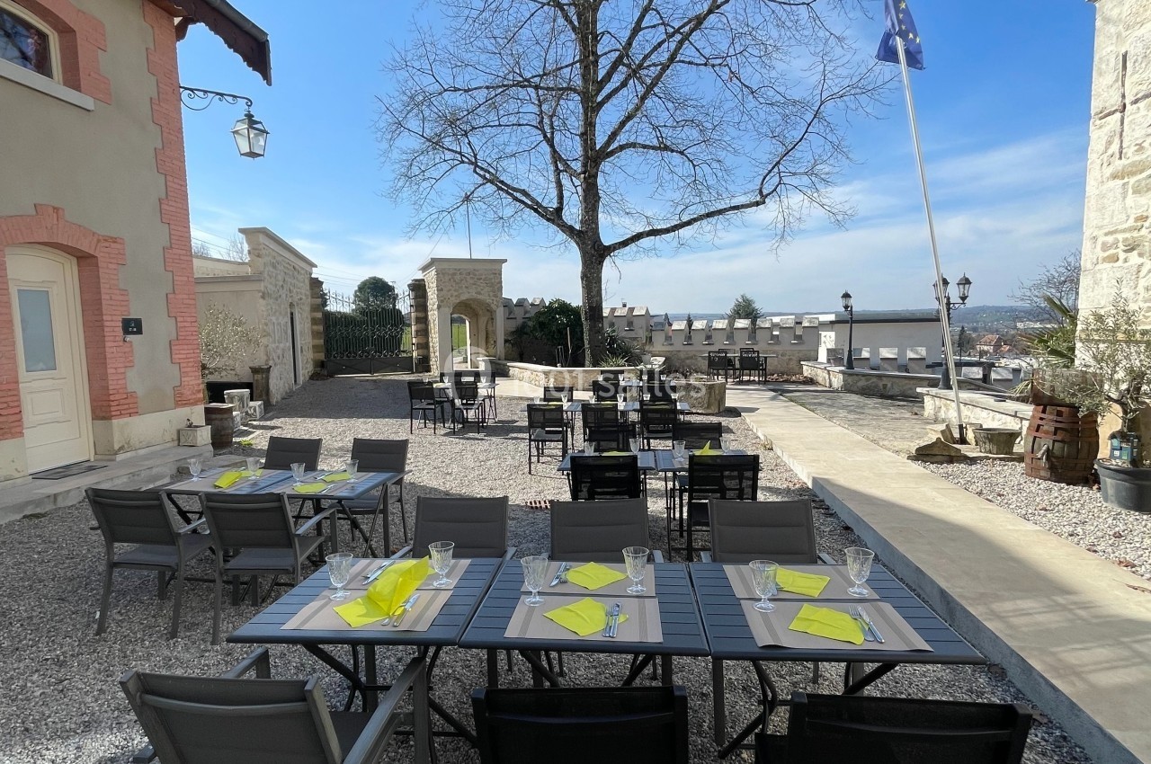 Terrasse extérieure avec tables dressées, chaises noires et vue dégagée sur un paysage, sous un grand arbre sans feuilles.
