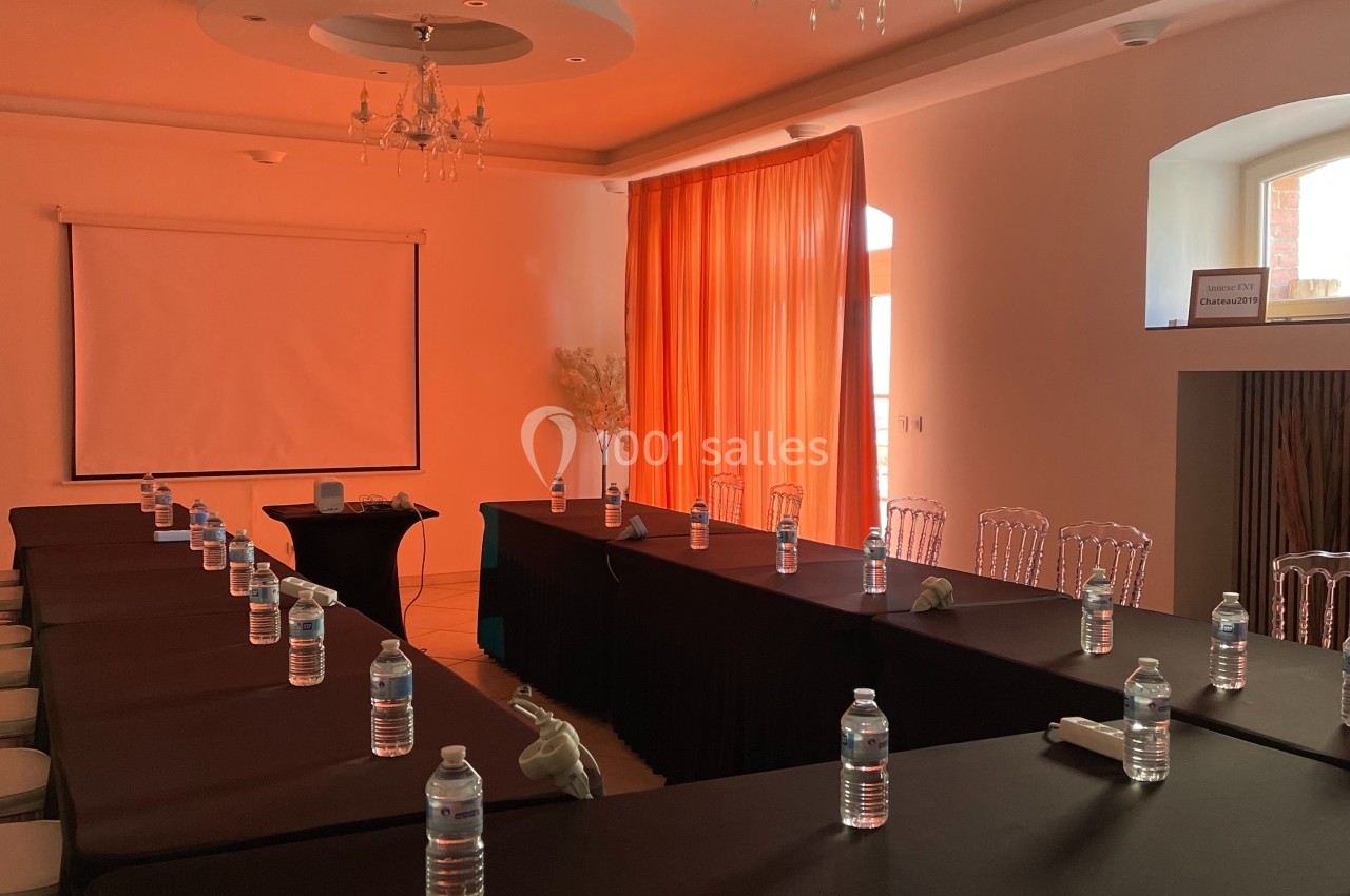 Salle de réunion avec tables en U, bouteilles d'eau alignées, rideaux orange et écran de projection en fond.