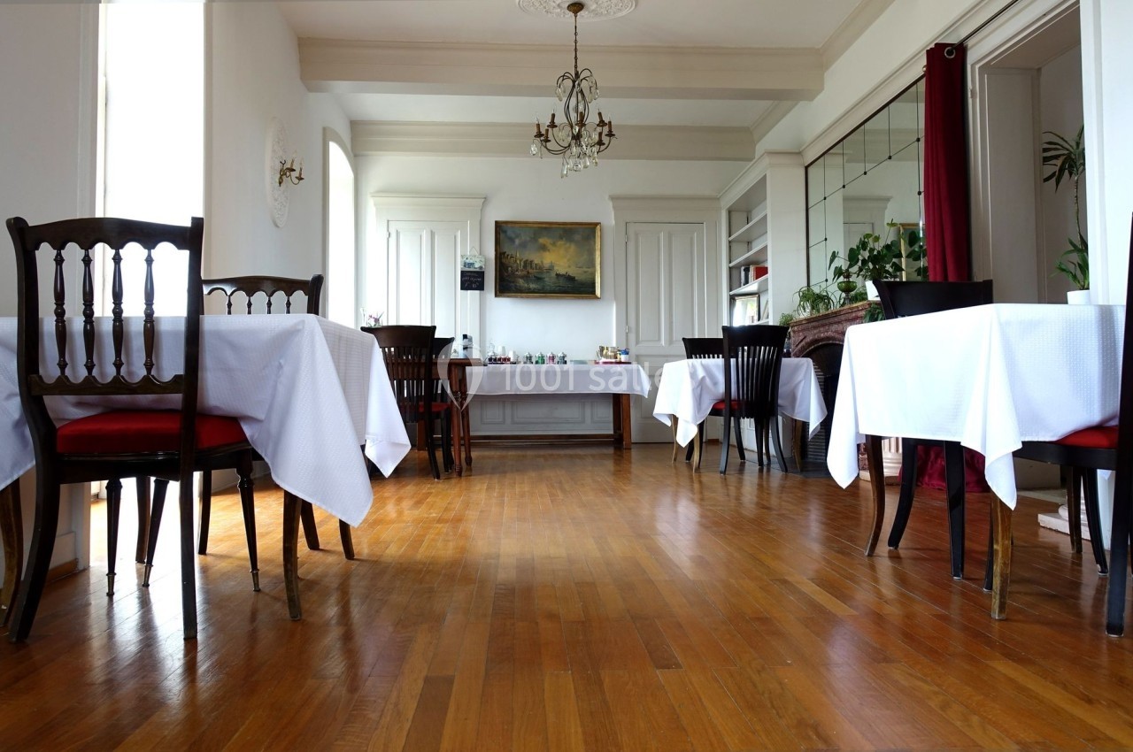 Salle à manger lumineuse avec parquet, tables dressées de nappes blanches, chaises en bois et décorations murales.