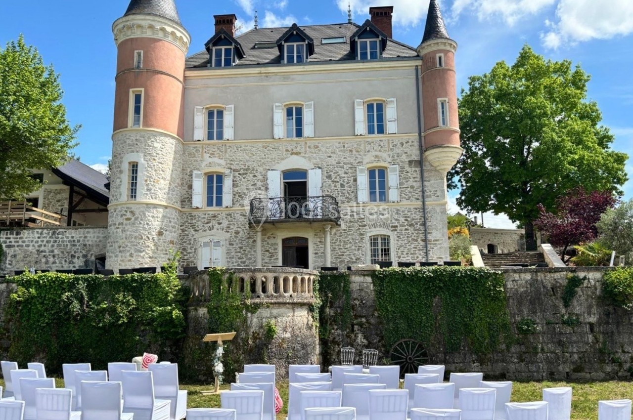 Façade d'un château en pierre avec deux tours, entouré de verdure, et des chaises blanches disposées en extérieur.