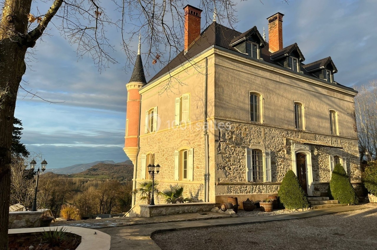 Façade d'un château en pierre avec une tour, éclairé par le soleil couchant, entouré d'arbres et d'un paysage vallonné.