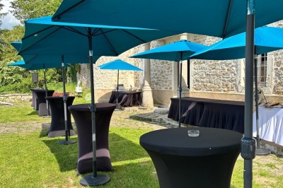 Façade d'un bâtiment en pierre avec une tour, des fenêtres blanches, une terrasse aménagée et un drapeau bleu.