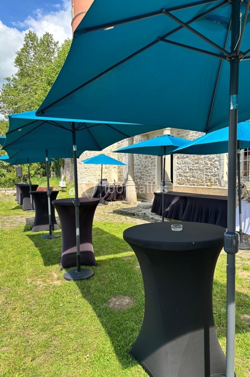 Parasols bleus et tables hautes noires disposés sur une pelouse devant un bâtiment en pierre par temps ensoleillé.