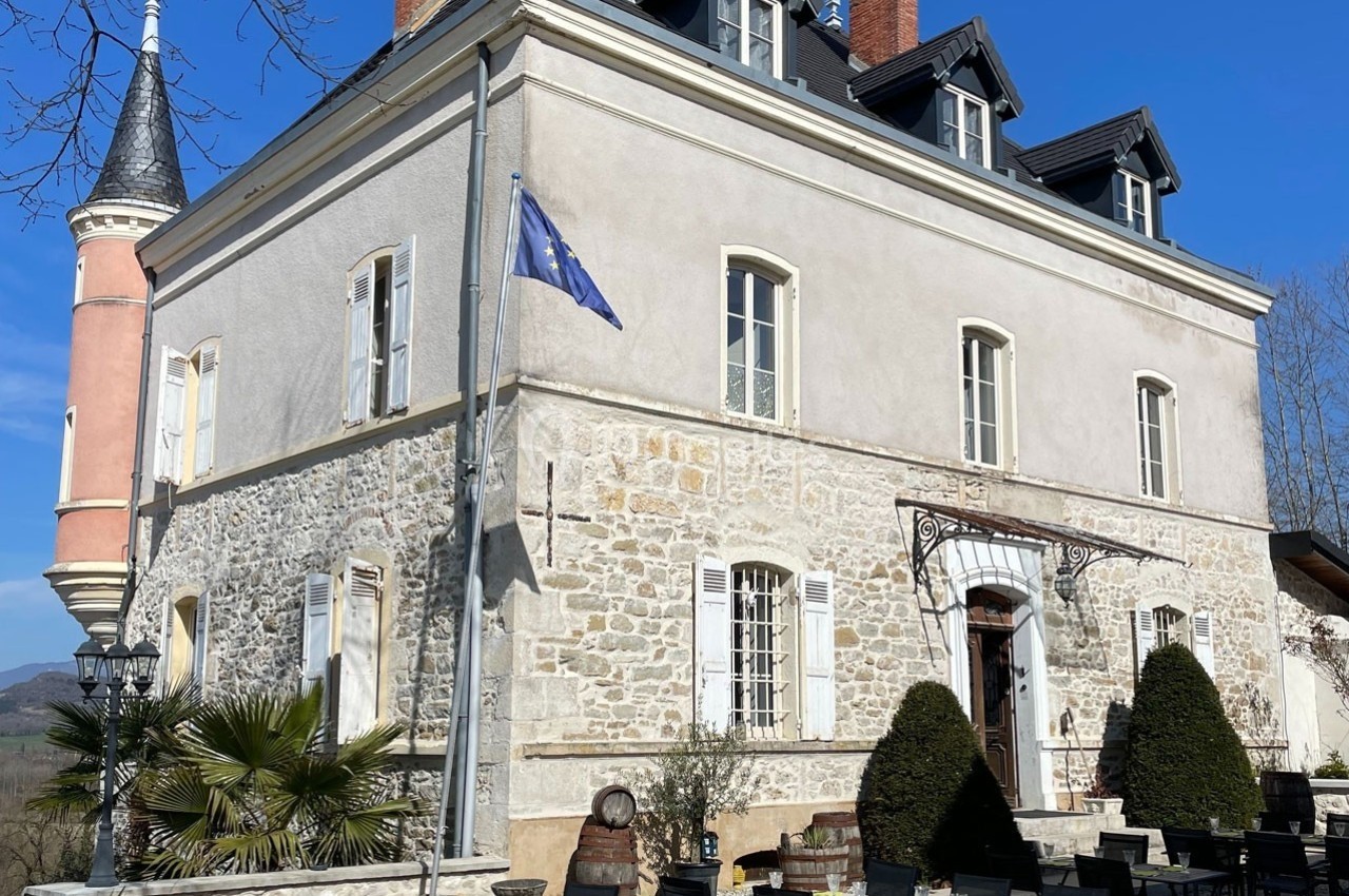 Façade d'un bâtiment en pierre avec une tour, des fenêtres blanches, une terrasse aménagée et un drapeau bleu.