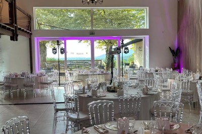 Façade d'un bâtiment en pierre avec une tour, des fenêtres blanches, une terrasse aménagée et un drapeau bleu.