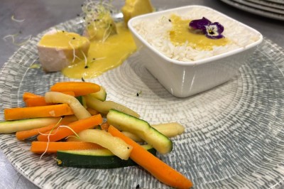 Façade d'un bâtiment en pierre avec une tour, des fenêtres blanches, une terrasse aménagée et un drapeau bleu.
