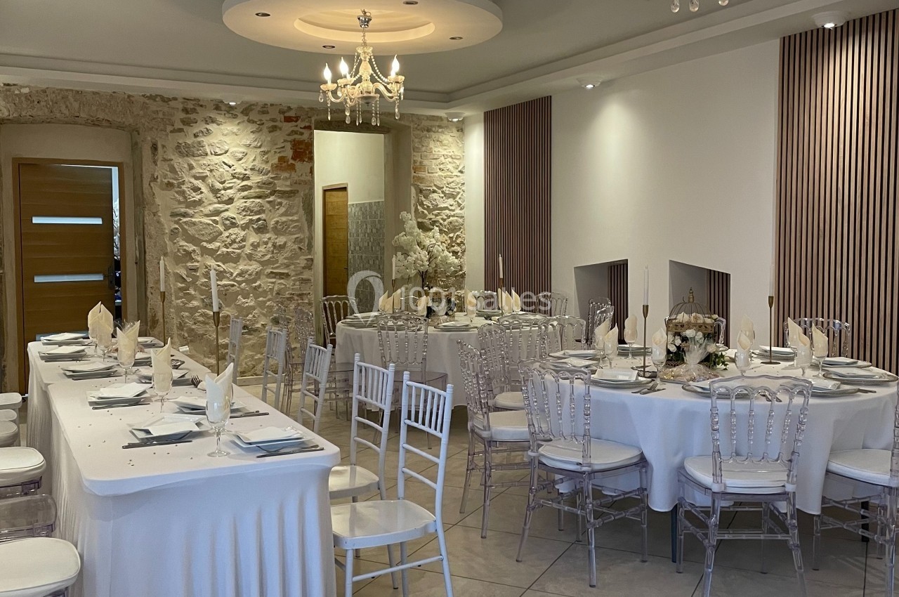 Salle de réception élégante avec tables décorées, chaises transparentes et murs en pierre apparente.