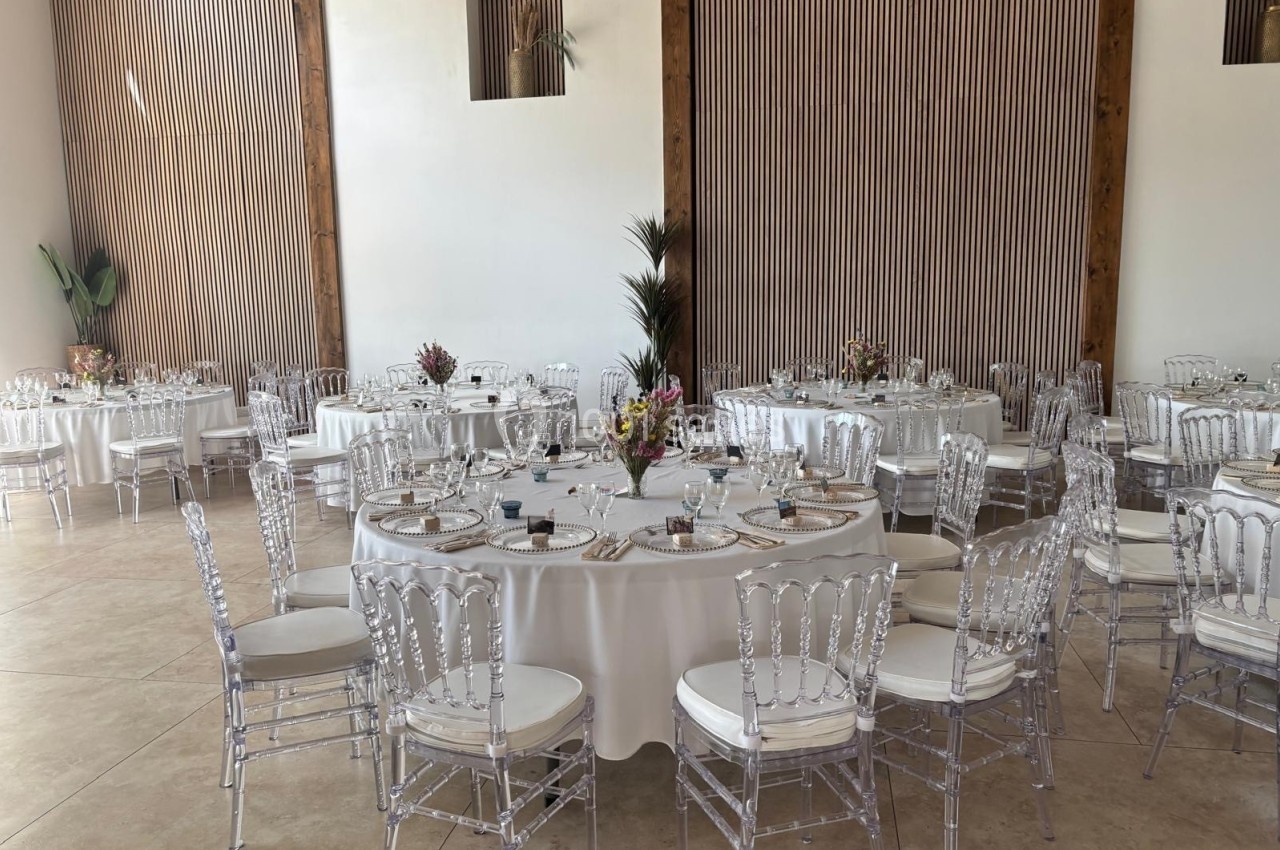 Salle de réception lumineuse avec des tables rondes dressées, nappes blanches et chaises transparentes.