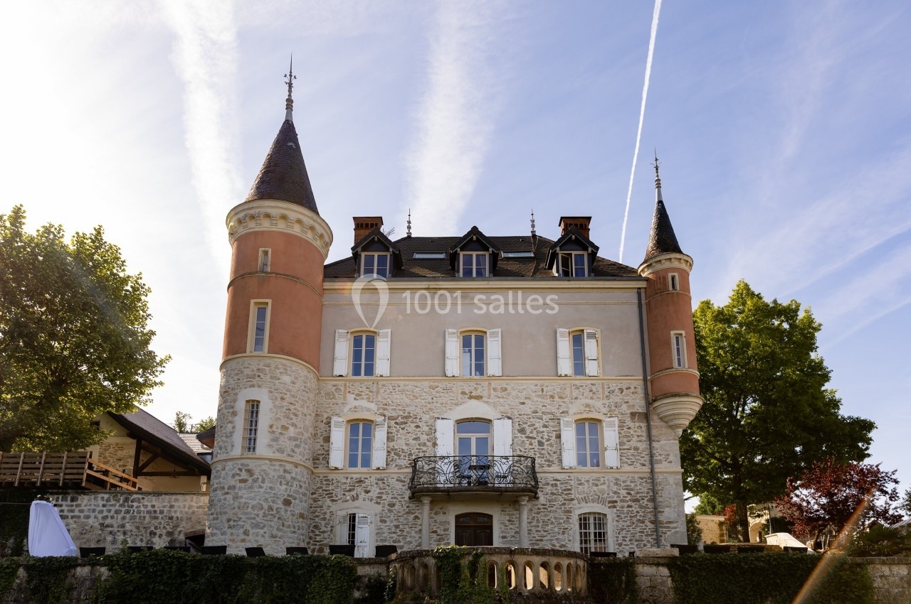 Façade d'un château en pierre avec deux tours rondes, entouré de verdure, sous un ciel bleu traversé de traînées d'avion.