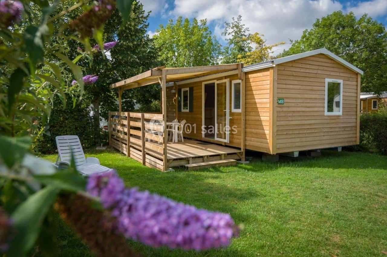Chalet en bois avec terrasse couverte, entouré de verdure et fleurs violettes au premier plan.