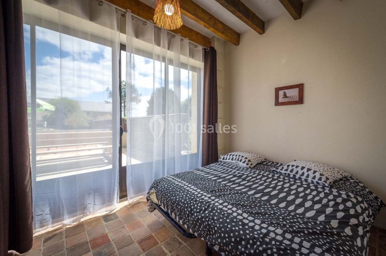 Chambre lumineuse avec un lit double, grandes baies vitrées donnant sur une terrasse ensoleillée et des rideaux blancs.