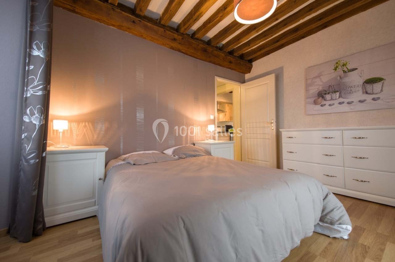 Chambre avec lit double, poutres apparentes, éclairage doux, murs décorés et mobilier blanc.