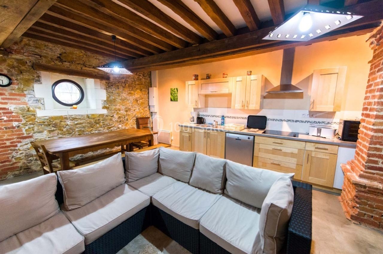 Cuisine ouverte rustique avec murs en pierre, poutres apparentes, table en bois et canapé en rotin.