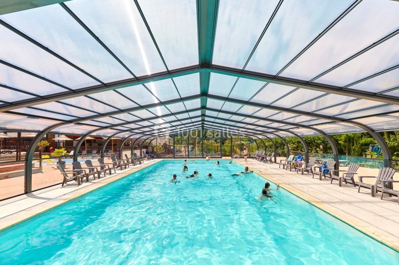 Piscine couverte avec des baigneurs, entourée de chaises longues et située dans un environnement ensoleillé.