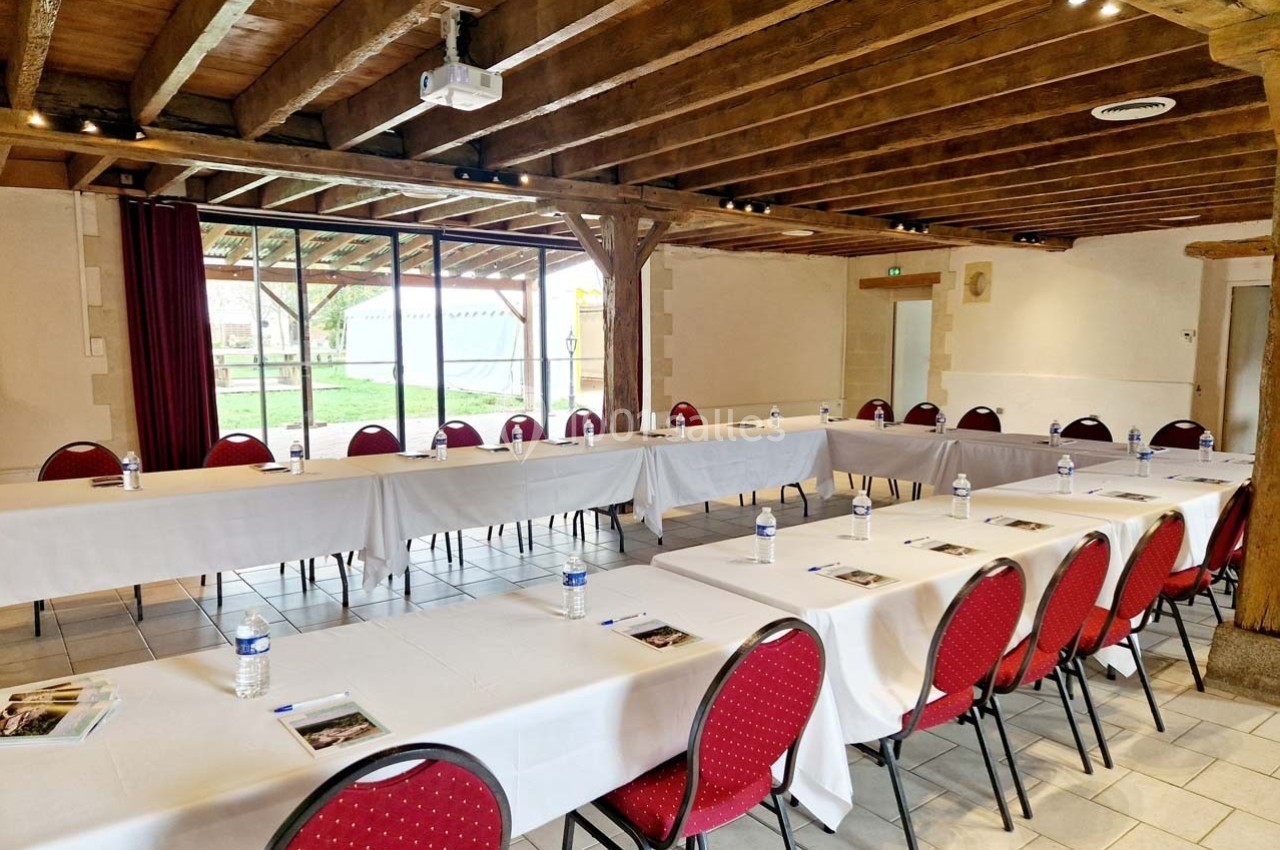 Salle de réunion lumineuse avec tables disposées en U, chaises rouges, bouteilles d'eau et documents sur chaque place.