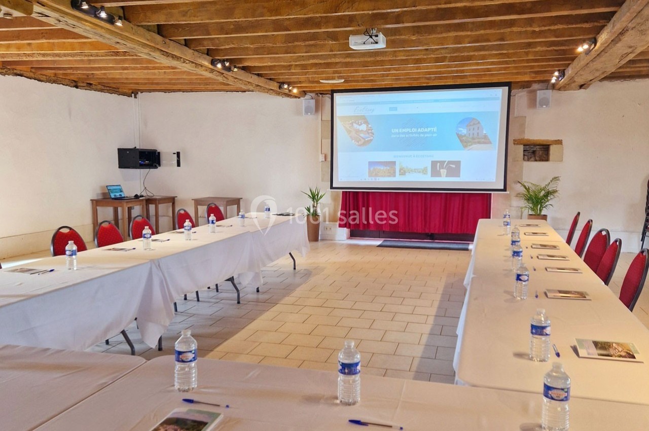 Salle de réunion lumineuse avec tables disposées en U, écran de projection, chaises rouges et poutres apparentes.