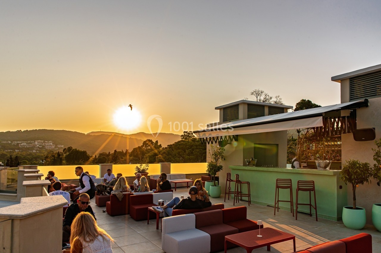 Terrasse ensoleillée avec des personnes assises, un bar moderne et une vue sur des collines au coucher du soleil.