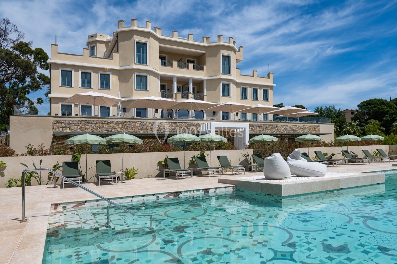Façade d'un hôtel beige avec terrasses, entouré de parasols et transats, devant une piscine extérieure ornée de motifs.