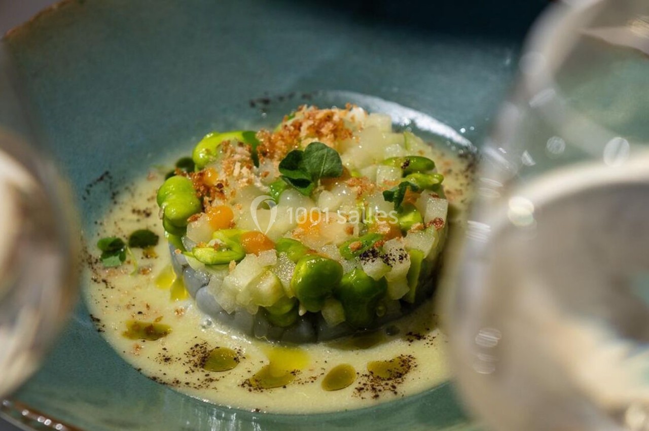 Tartare de légumes colorés servi dans une assiette en céramique bleue, entouré de deux verres flous au premier plan.
