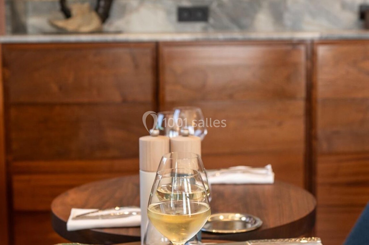 Un verre de vin blanc posé sur une table en bois, avec des couverts et des verres en arrière-plan dans un décor chaleureux.