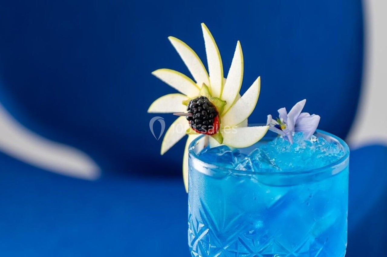 Verre de cocktail bleu garni de tranches de pomme, d'une mûre et d'une fleur, posé devant un fond bleu.