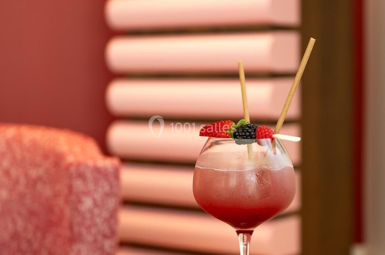Verre de cocktail rouge garni de fruits rouges et pailles, posé sur une table devant un décor intérieur rose.