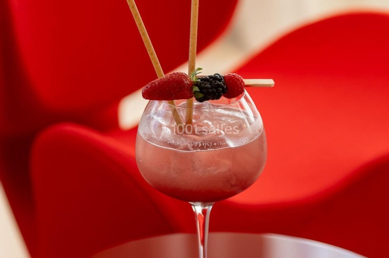 Verre de cocktail rouge garni de fraises et de mûres, posé sur une table devant un fauteuil rouge.