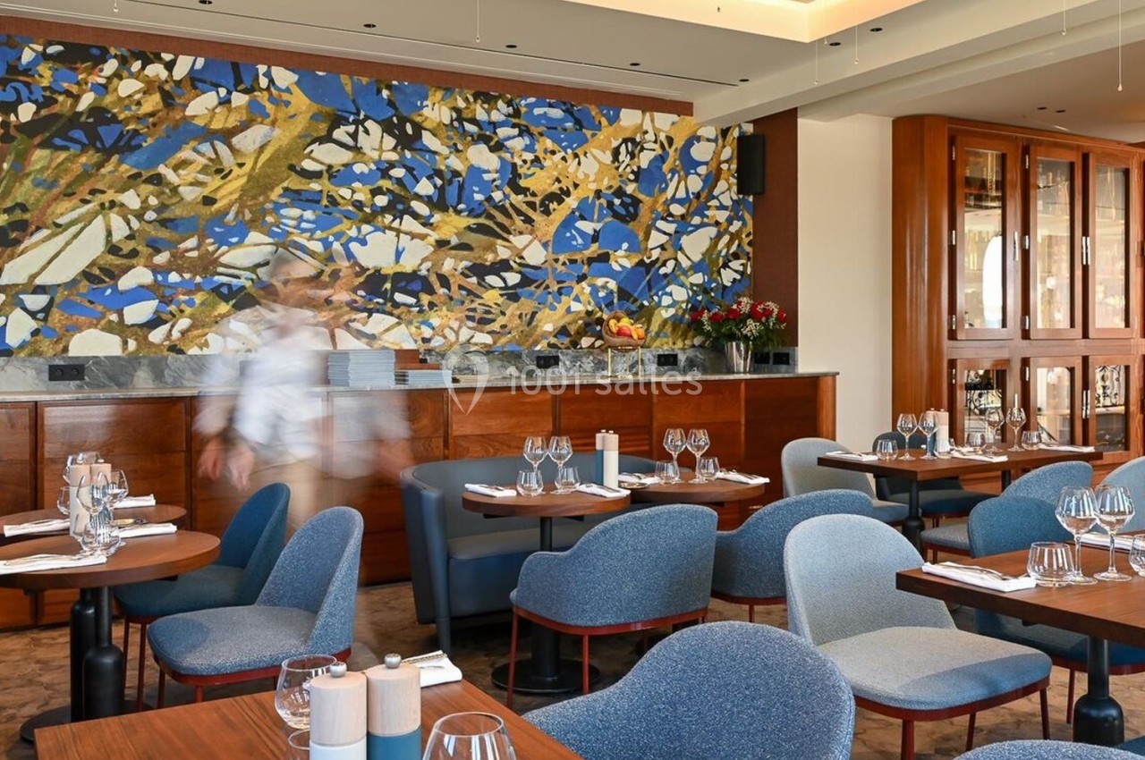 Salle de restaurant élégante avec tables dressées, chaises bleues et grande fresque colorée sur le mur du fond.