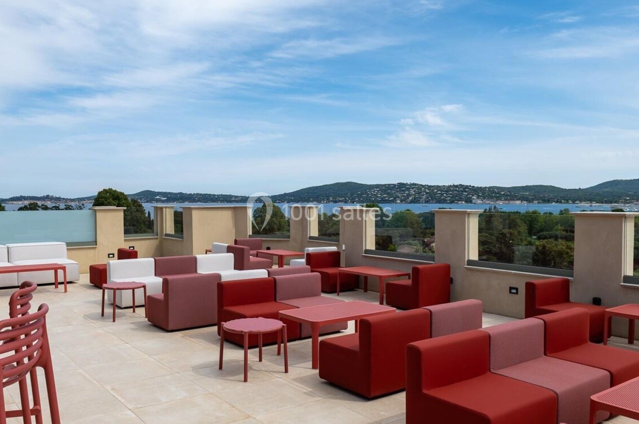 Terrasse avec mobilier rouge et blanc, offrant une vue panoramique sur des collines, un lac et un ciel dégagé.