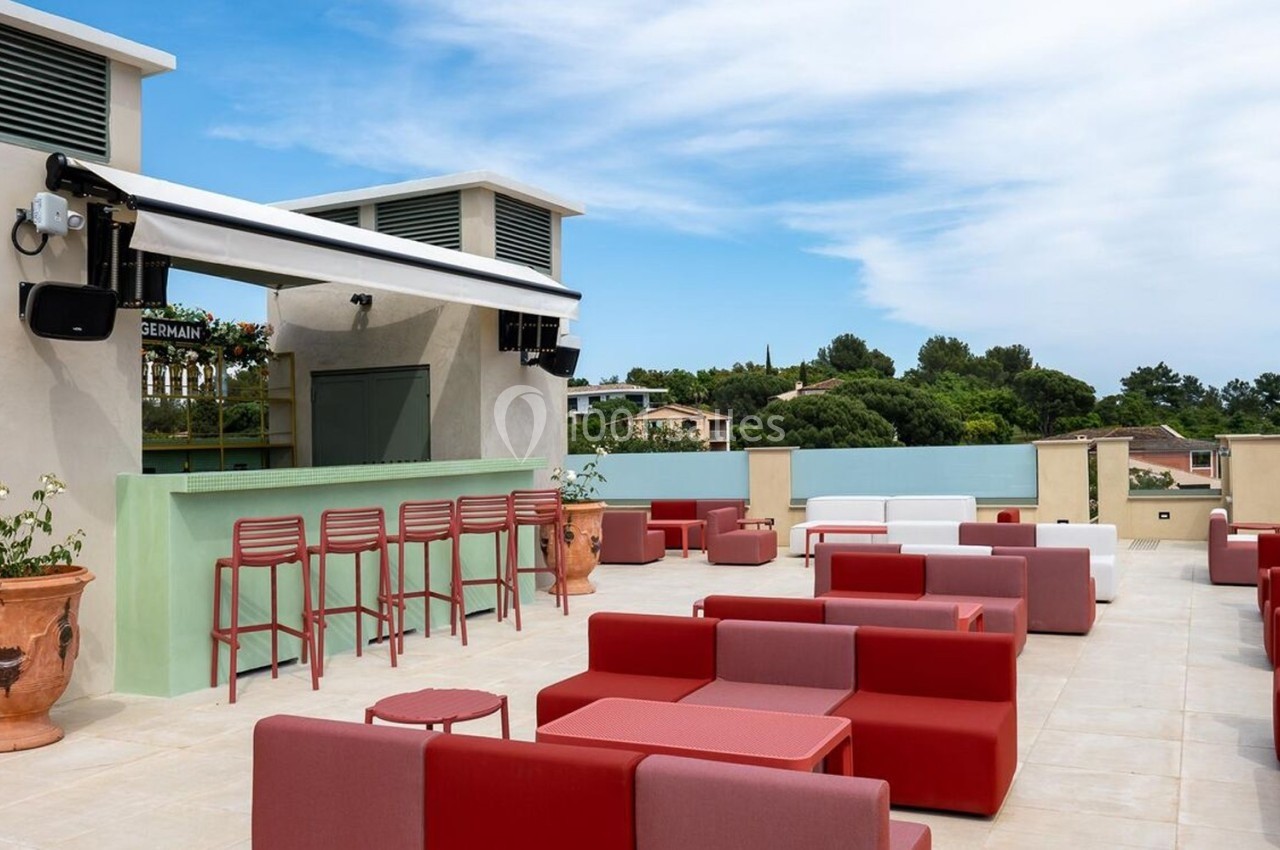 Terrasse extérieure avec bar, chaises hautes rouges, tables et fauteuils colorés, entourée de verdure et ciel dégagé.