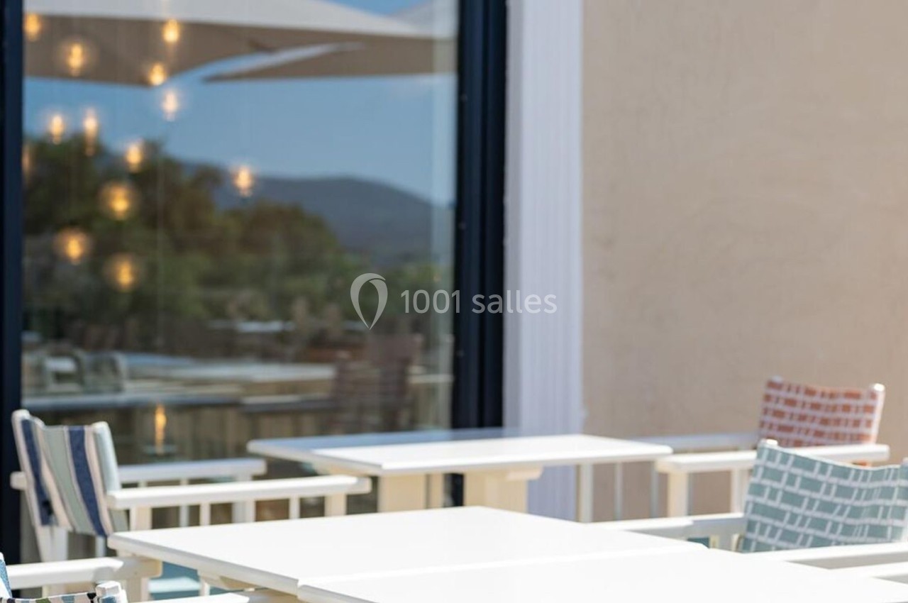Tables et chaises blanches disposées sur une terrasse ensoleillée, avec un reflet de paysage dans une baie vitrée.