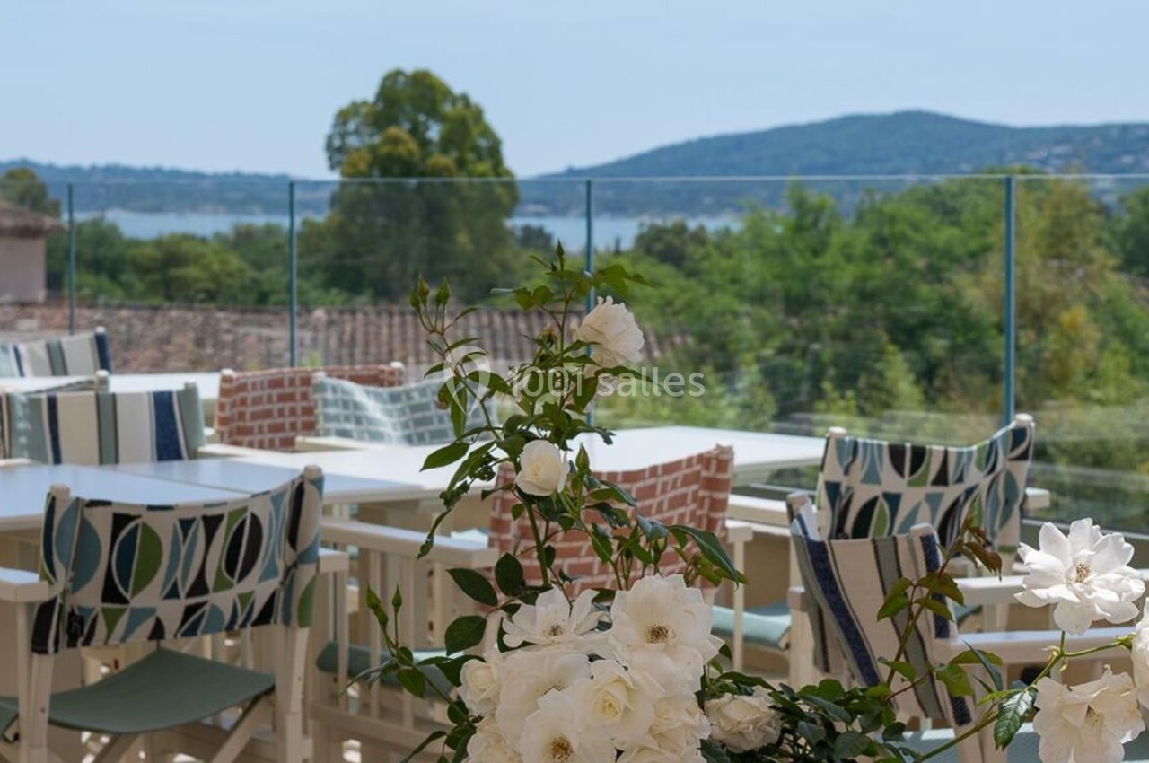 Terrasse avec tables, chaises et fleurs blanches, offrant une vue sur des collines verdoyantes et un plan d'eau.