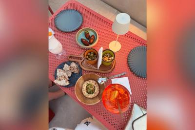 Table rouge avec assiettes, pain, olives, tomates, houmous, boisson orange et lampe, en extérieur.