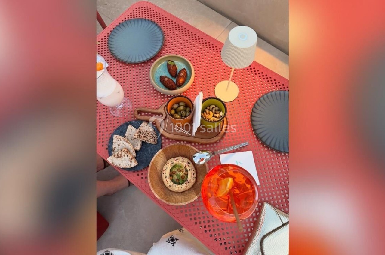 Table rouge avec assiettes, pain, olives, tomates, houmous, boisson orange et lampe, en extérieur.