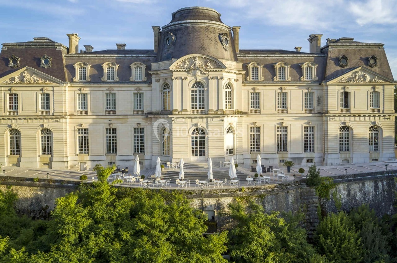 Façade d'un château historique avec terrasses et parasols, entouré de verdure sous un ciel dégagé.