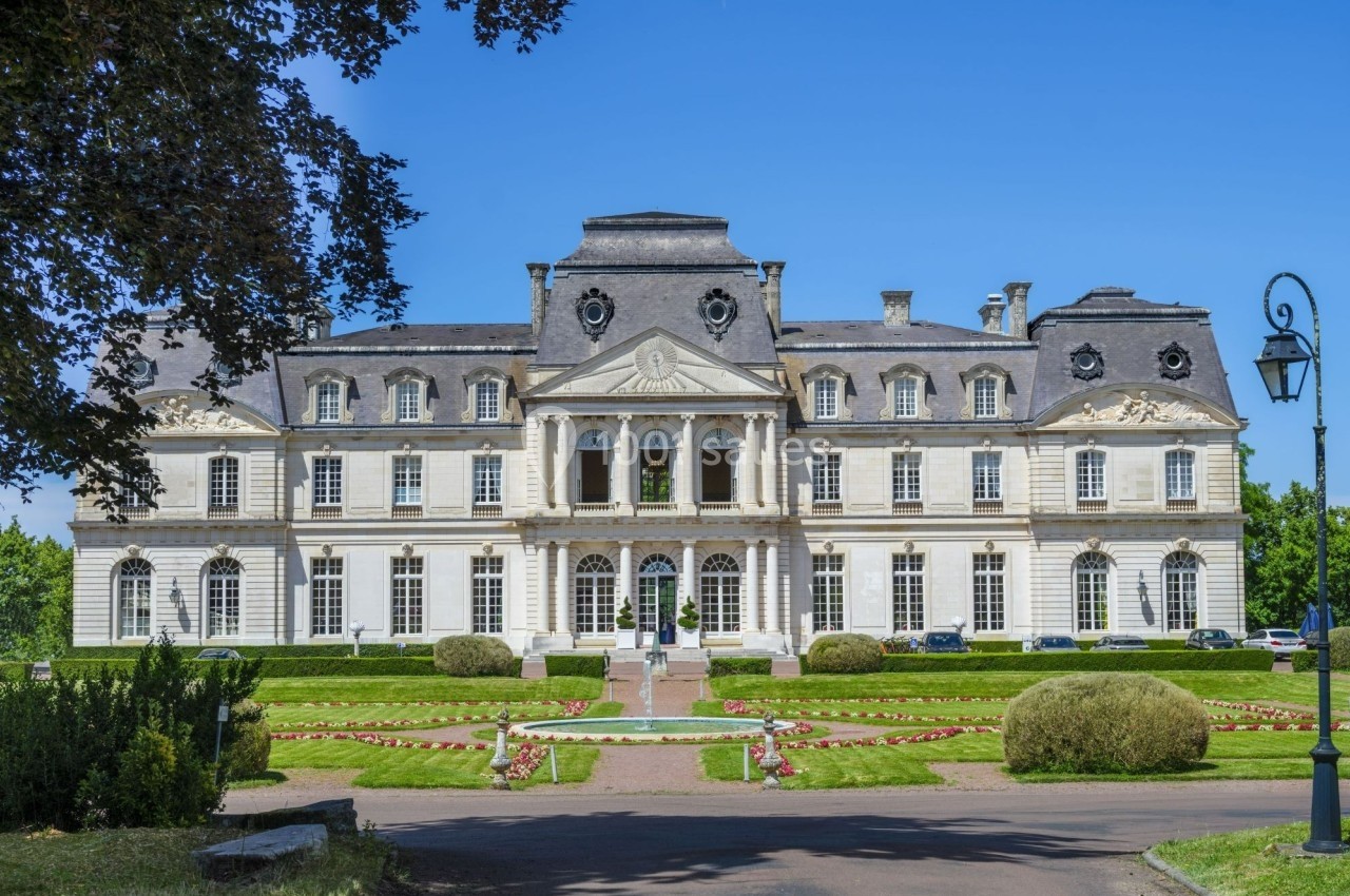 Façade d'un grand bâtiment historique en pierre avec un jardin aménagé et un ciel bleu en arrière-plan.