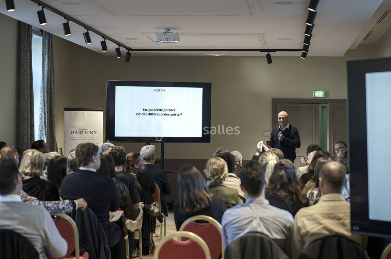 Un conférencier s'adresse à un public assis dans une salle équipée d'écrans et de matériel audiovisuel.