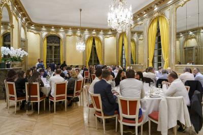 Salle à manger élégante avec table dressée, chaises rouges, murs dorés, lustres et grandes fenêtres avec rideaux jaunes.