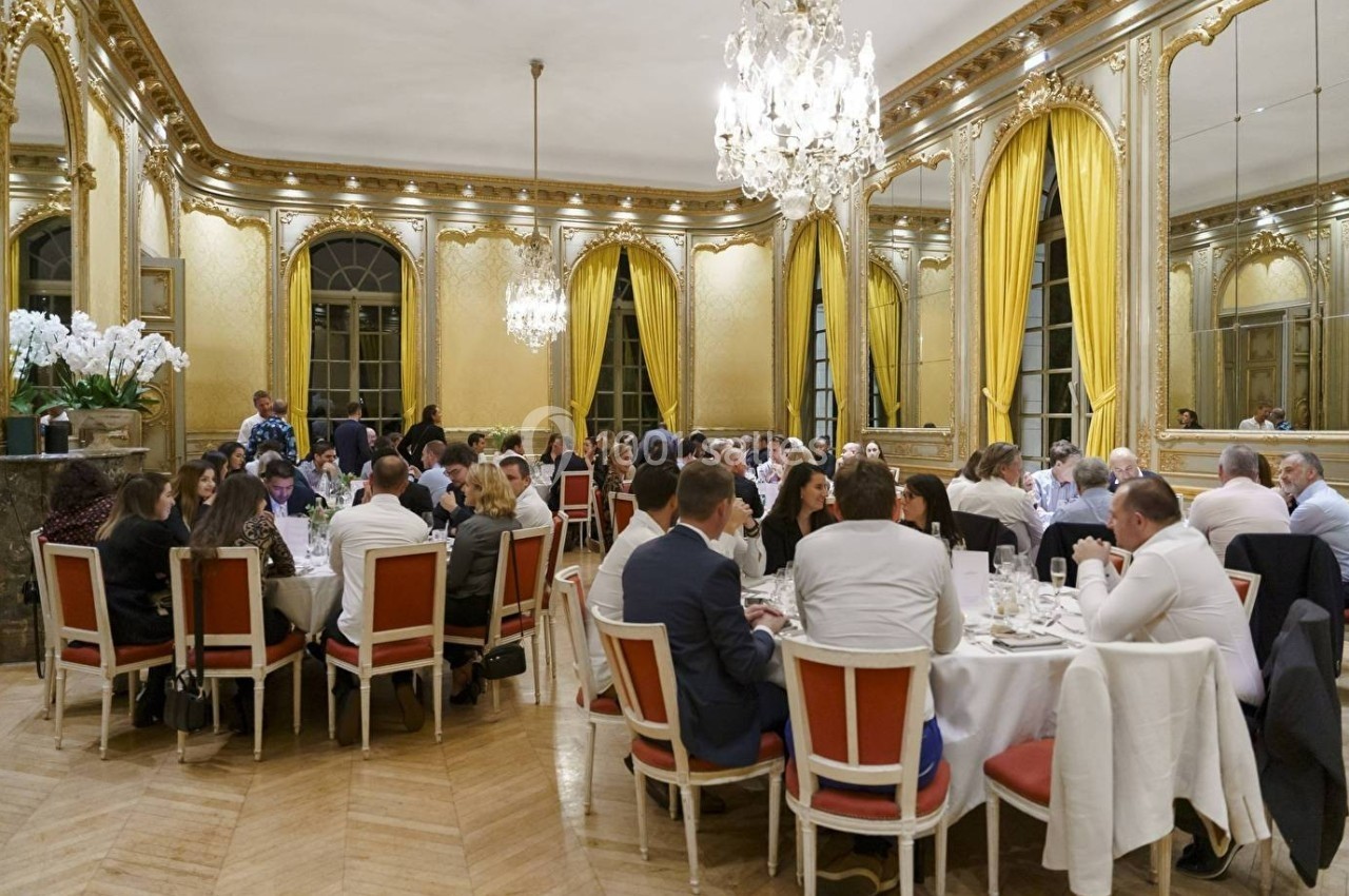 Des convives assis autour de tables rondes dans une salle élégante avec miroirs, lustres et rideaux dorés.