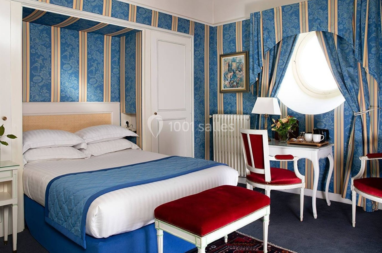 Chambre lumineuse avec lit double, murs à rayures bleues, mobilier classique blanc et rouge, et fenêtre ronde.