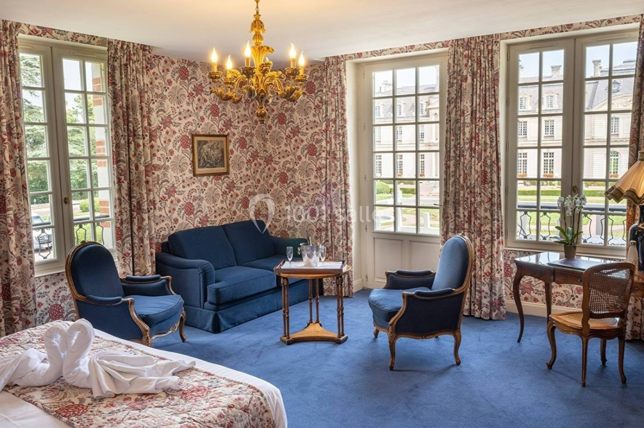 Chambre lumineuse avec mobilier classique, papier peint fleuri, lit, coin salon et vue sur un bâtiment historique.