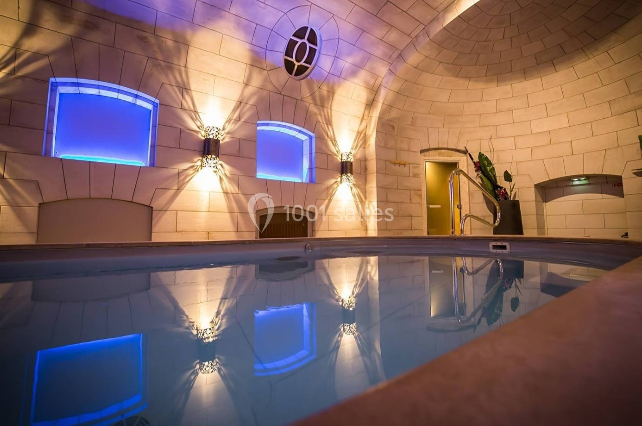 Piscine intérieure éclairée par des lumières bleues, entourée de murs en pierre et de plantes décoratives.