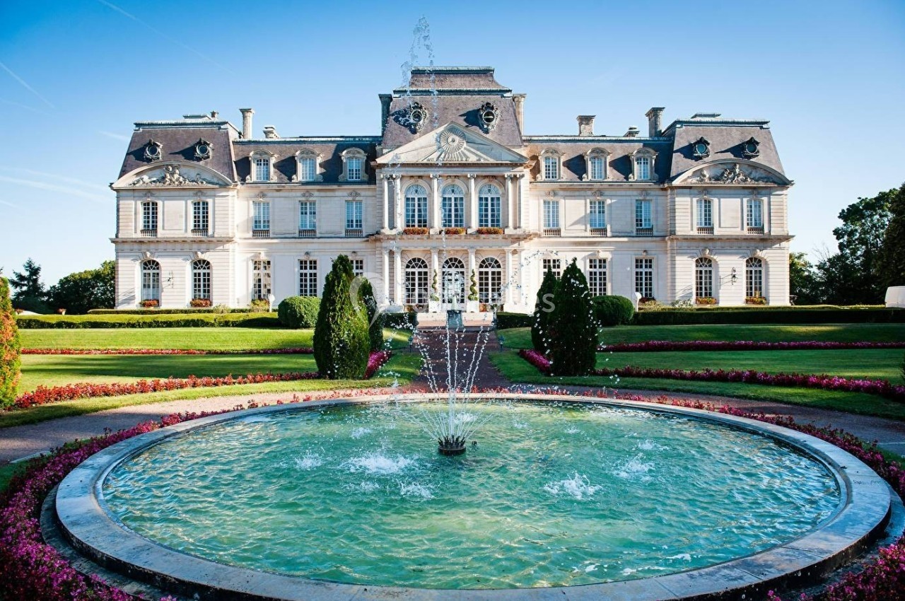 Façade d'un grand château entouré de jardins paysagers avec une fontaine circulaire au premier plan.