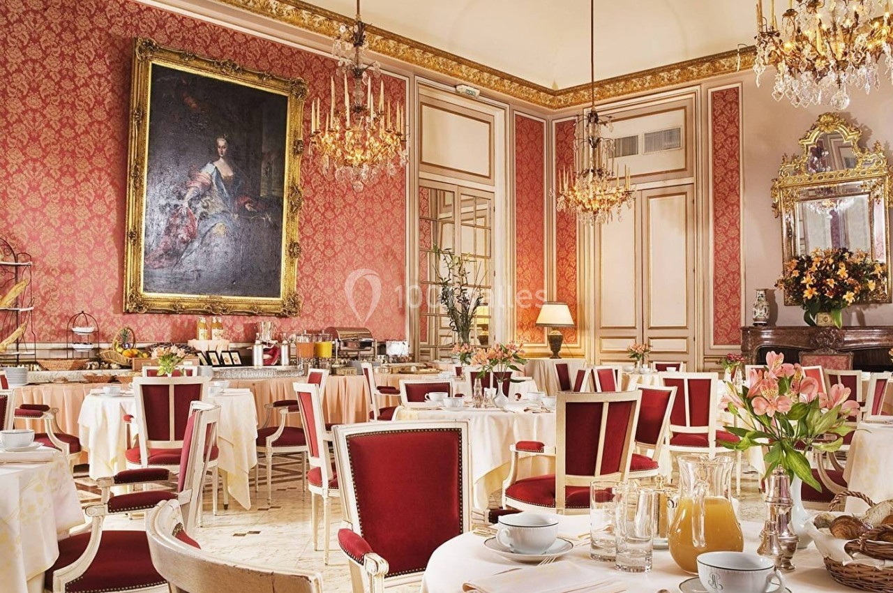 Salle de restaurant élégante avec murs rouges ornés, lustres dorés, tables dressées et grand tableau ancien.